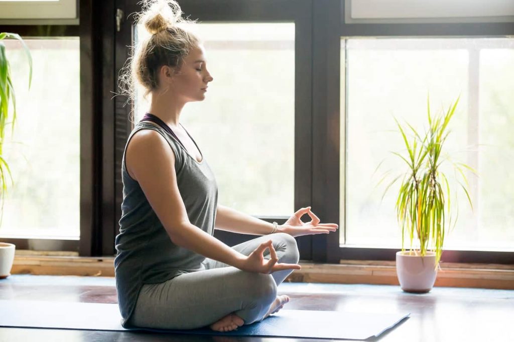 young blonde woman cross legged on yoga mat meditating beside calming indoor plants